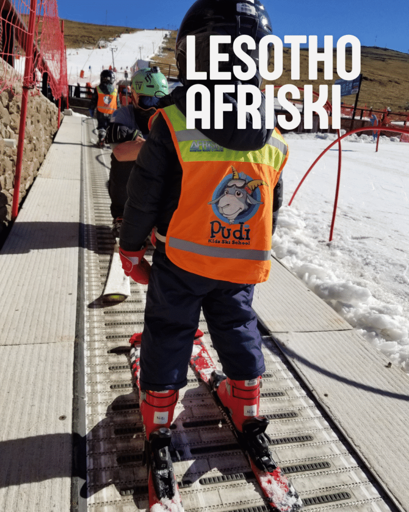 Three-year-old on magic carpet ski lift at Lesotho Afriski Resort with Pudi orange ski school vest and long ski run in background surrounded by dry arid grasslands showcasing it wasn't quite cold enough for real snow.